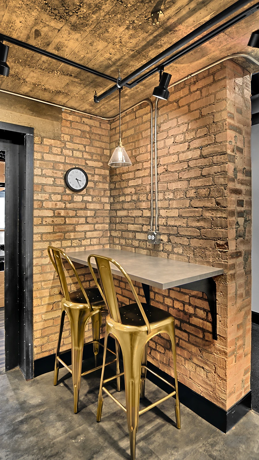 Office Interior Industrial brick-walled corner with a metal table and two gold metal bar stools under exposed piping and a hanging lamp.