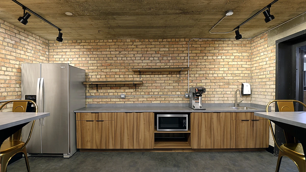 Office Interior Industrial kitchen with brick walls, stainless refrigerator, wooden cabinets, microwave, coffee machine, sink, and lighting.