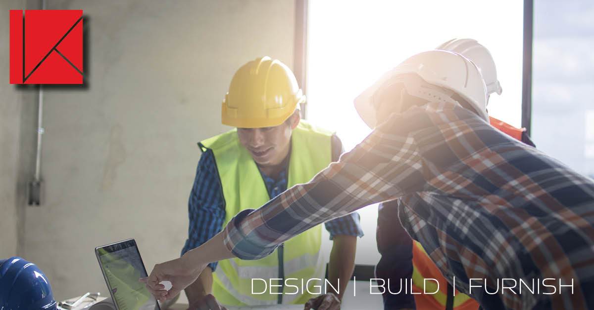 Two men in hard hats discuss office construction with a laptop nearby