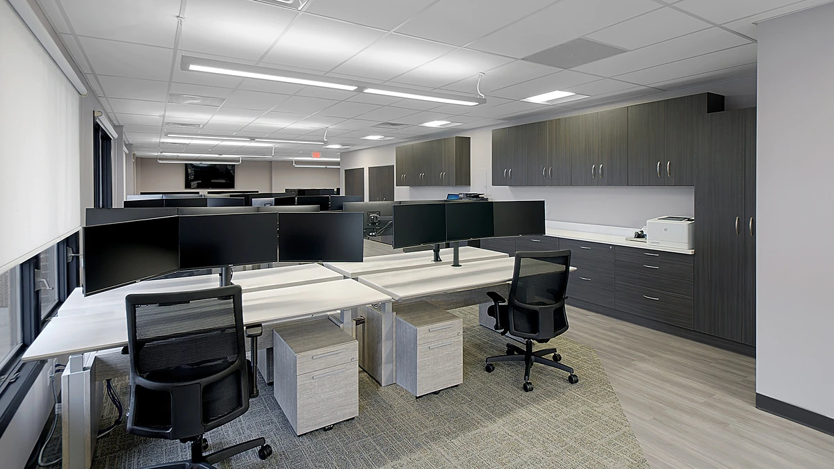 Office Interior Open-plan office with row of desks, dual-monitor setups, black chairs, grey cabinetry, and a large screen in the distance.
