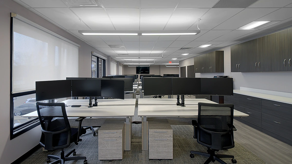 Office Interior Open-plan office with long rows of dual-monitor desks, black ergonomic chairs, and gray cabinets along the walls.