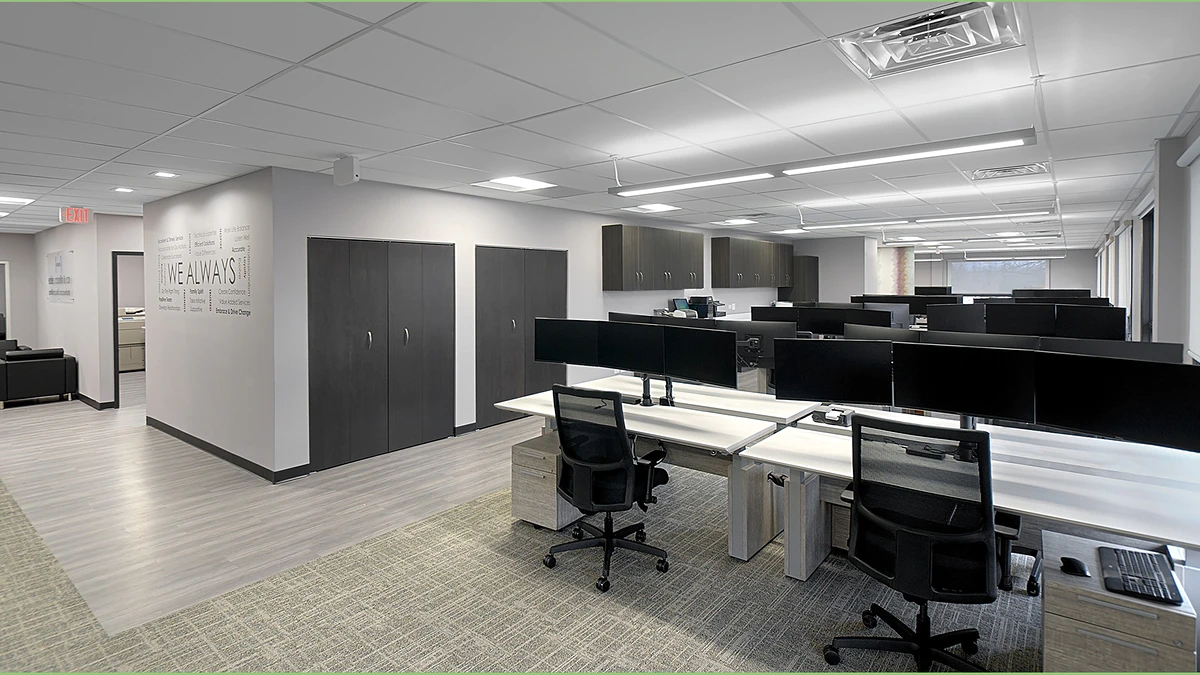Office Interior Open-plan office with rows of desks, dual monitors, black chairs, gray cabinets, and bright ceiling lights.