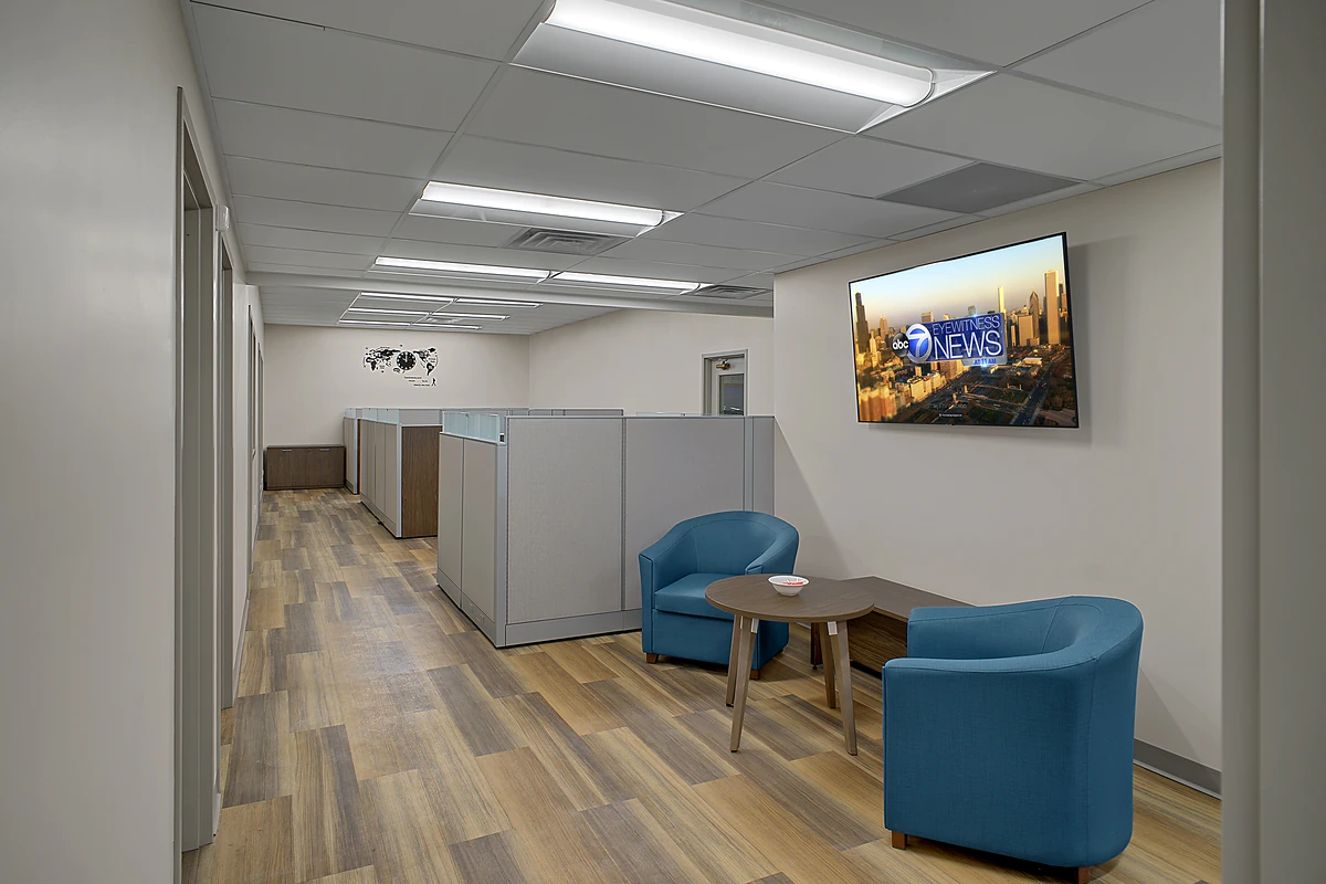 Office Interior Modern office lounge with cubicle partitions, two blue chairs around a round table, wall TV showing ABC7 News.