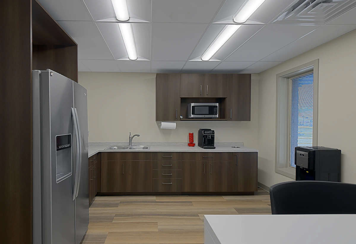 Office Interior A modern office kitchenette with a stainless steel fridge, wooden cabinets, sink, microwave, coffee maker, and window.