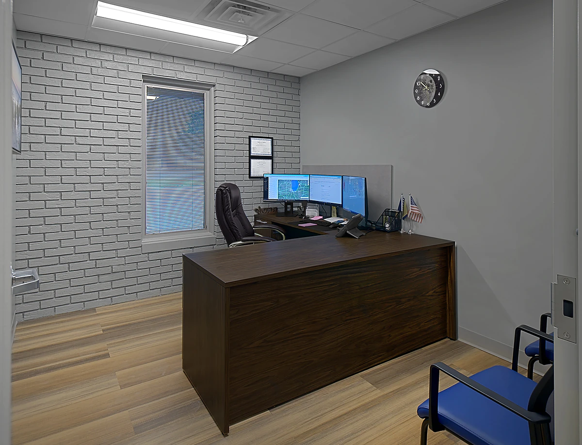 Office Interior Office workspace with dark wood desk, three monitors, brick wall, window with blinds, clock, and small flags.